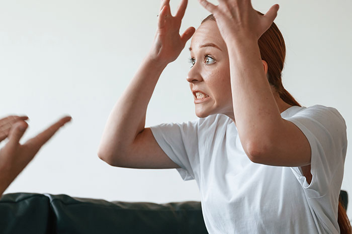 Frustrated young woman raising hands arguing with partner on couch, showing tension in relationship situation.
