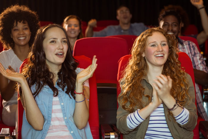 Two women happily clapping in a theater, illustrating a lady’s unpunctual friend painting nails before event.