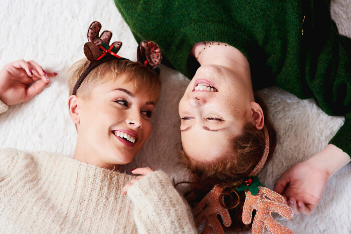Two women wearing holiday reindeer headbands lying down smiling, relating to partner holiday gift relationship insights. Two women wearing holiday reindeer headbands lying down smiling, relating to partner holiday gift relationship insights.