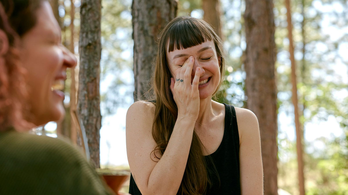 Two women laughing outdoors in a forest setting illustrating harmless but toxic habits discussion.