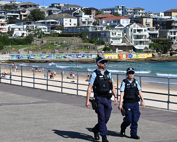 Two police officers walking near Bondi Beach with beachgoers and residential buildings in the background on a sunny day Two police officers walking near Bondi Beach with beachgoers and residential buildings in the background on a sunny day