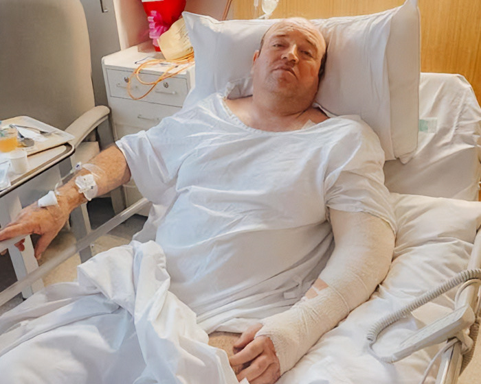 Man identified as hero cop who stopped Bondi Beach attacker, lying in hospital bed with bandaged arm and IV in hand.