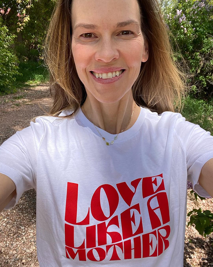 Hilary Swank outdoors smiling, wearing a white shirt with red text, related to apology and mom of terminally ill kids controversy.