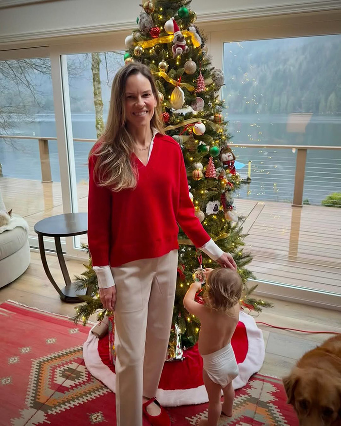 Woman in red sweater stands by Christmas tree with toddler decorating an ornament, relating to Hilary Swank and terminally ill kids. Woman in red sweater stands by Christmas tree with toddler decorating an ornament, relating to Hilary Swank and terminally ill kids.