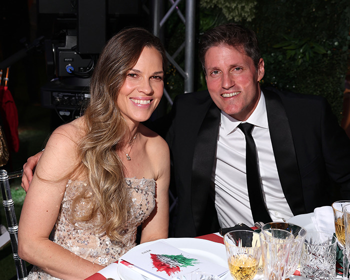 Hilary Swank smiles seated next to a man in a black suit at a formal dinner event outdoors. Hilary Swank smiles seated next to a man in a black suit at a formal dinner event outdoors.