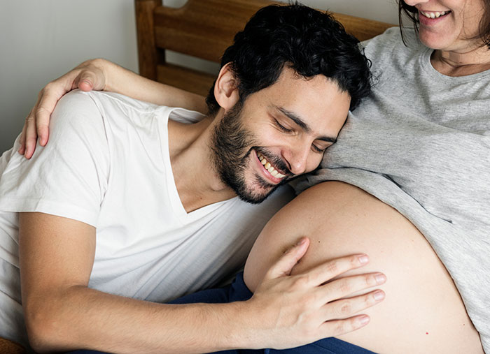 Couple expecting newborn, man smiling while resting head on pregnant woman's belly, tension hinted by body language.