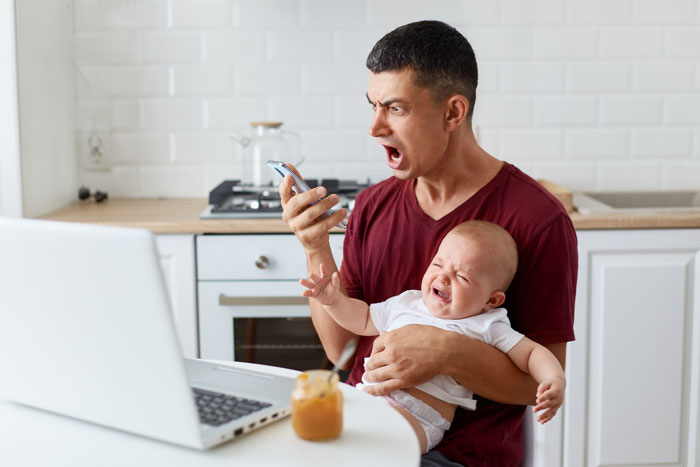Frustrated dad holding crying baby while looking at phone, highlighting stay-at-home-mom chores and parenting stress.