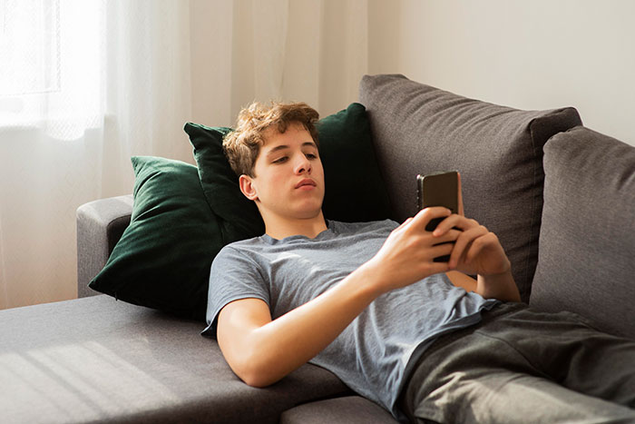 Teen boy lying on sofa using smartphone, illustrating dating advice from dad straight out of 1955 in a modern home.