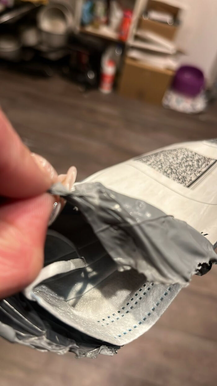Close-up of a hand holding a damaged grey plastic package with a face mask inside in a messy room setting.