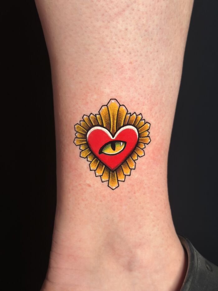 Tattoo of a red heart with a cat’s eye in the center, a colorful pet tribute inked on inner ankle skin.