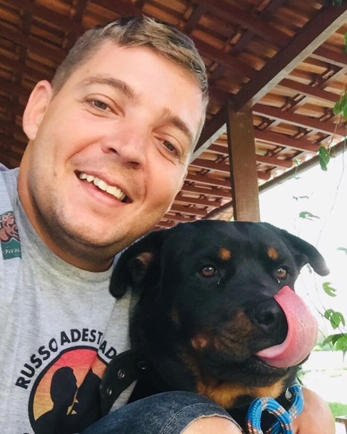 Smiling man taking a close selfie with a large black and brown dog wearing a blue collar under a wooden roof. Smiling man taking a close selfie with a large black and brown dog wearing a blue collar under a wooden roof.