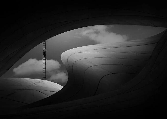 Minimalism art photo of a person climbing a ladder between smooth curved structures under a cloudy sky.