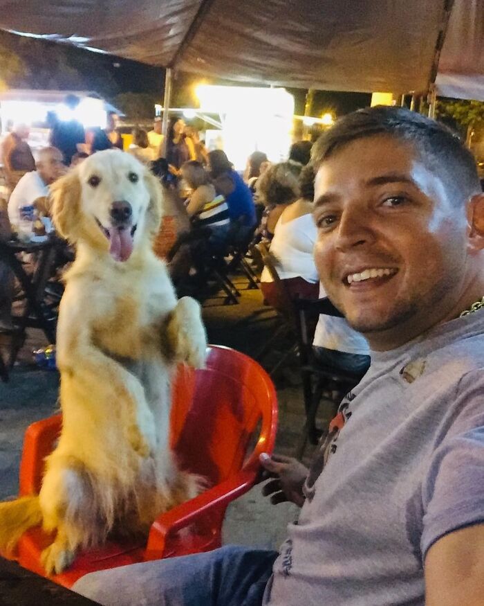 Man smiling and sitting next to a golden retriever dog in an outdoor dining area at night with people in the background Man smiling and sitting next to a golden retriever dog in an outdoor dining area at night with people in the background