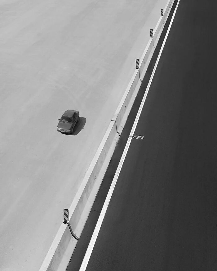 Black and white minimalism photo of a single car on a wide road beside a contrasting dark highway and barrier wall.