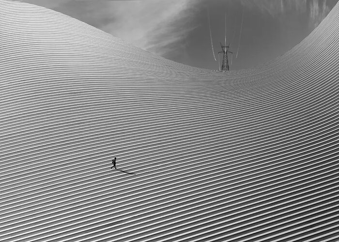 Minimalism photography showing a lone figure walking on textured, striped architectural surface with dramatic lighting and shadows.