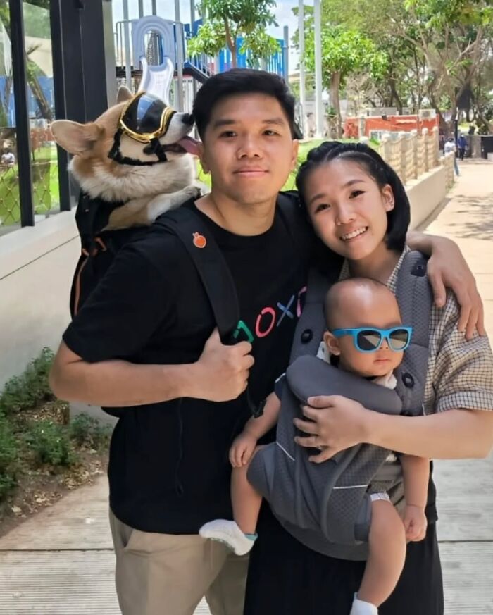 Man carrying dog in backpack wearing sunglasses standing with woman holding baby in carrier on city sidewalk