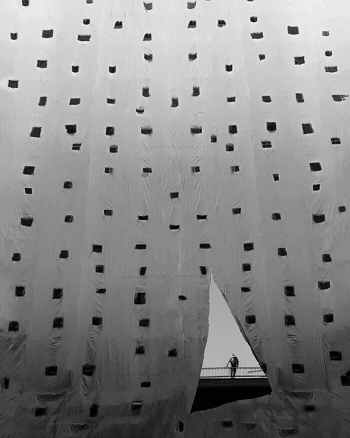 Minimalist black and white photo featuring a large fabric with cutouts and a solitary person framed in a triangular opening.