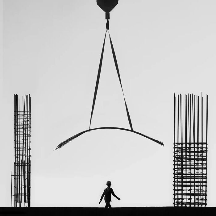 Minimalism art photograph showing a worker walking beneath a piece of steel suspended by a crane with construction materials on sides.