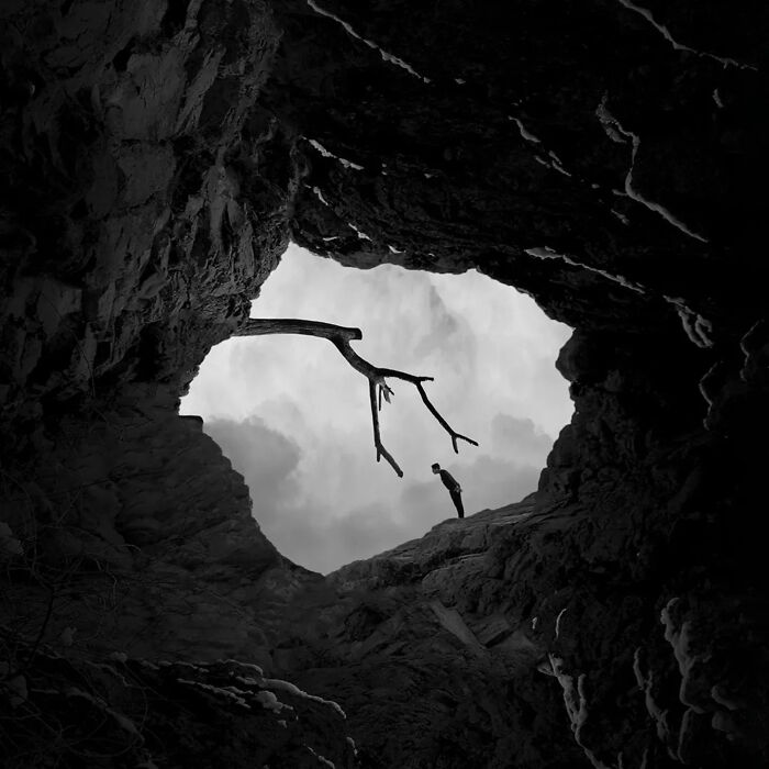 Black and white minimalism image showing a person standing inside a rocky cave looking at a bare tree branch against the sky.