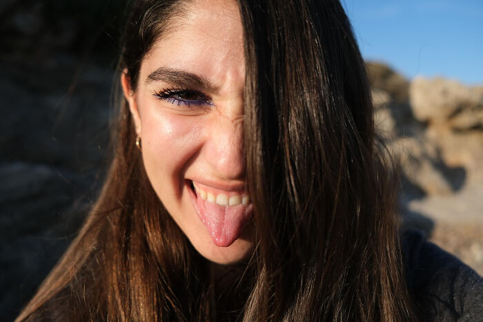 Young woman showing playful expression with tongue out, highlighting fascinating human body features in natural sunlight outdoors.