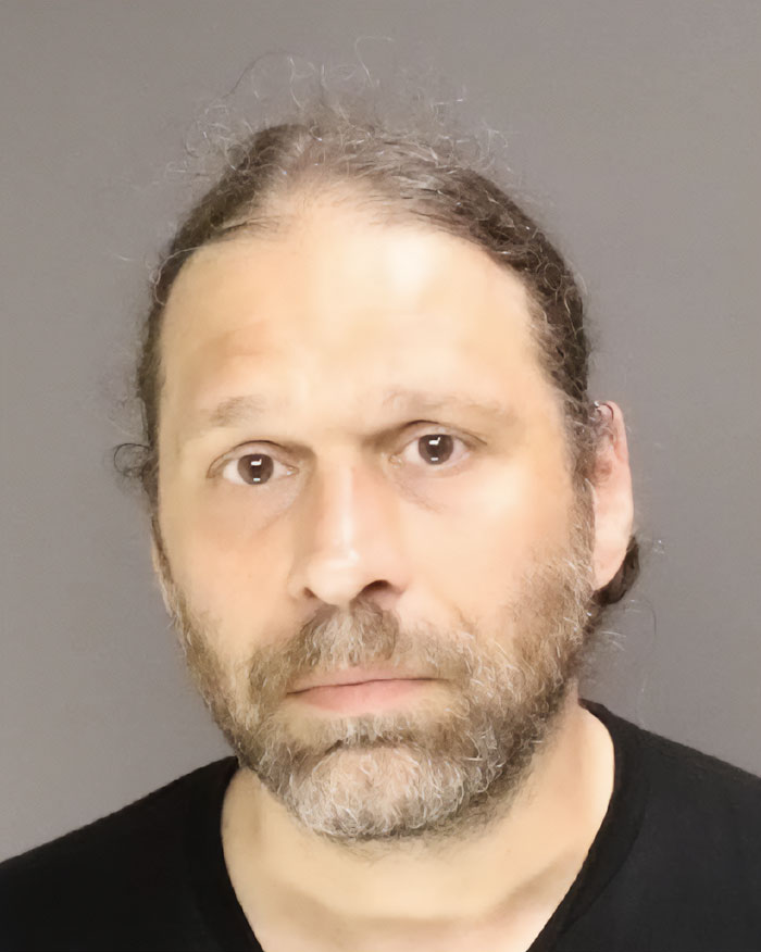 Man with long hair and beard, wearing a black shirt, shown in a close-up mugshot related to NYU shove victim viral video. Man with long hair and beard, wearing a black shirt, shown in a close-up mugshot related to NYU shove victim viral video.