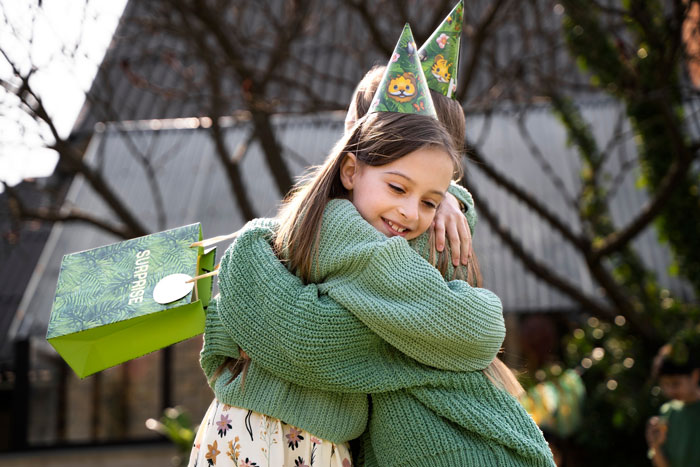 Jehovah's Witness mom hugging child at birthday party wearing party hats and holding a green surprise gift bag outdoors.