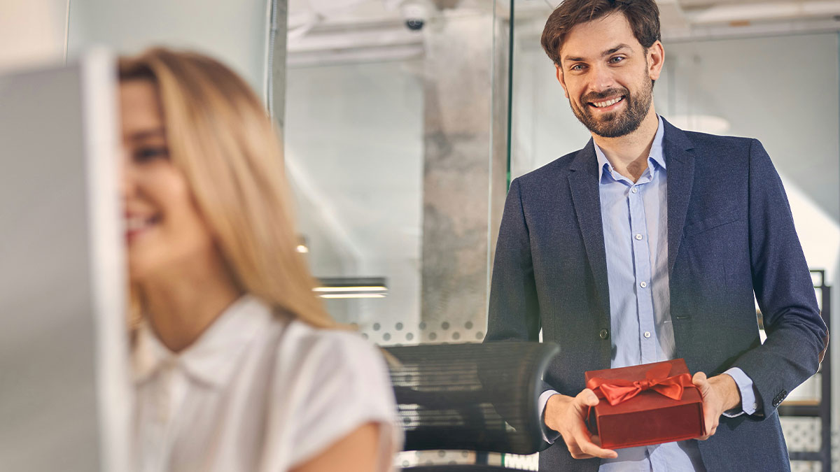 Smiling man in business attire holding a gift box behind a blurred woman in an office setting, job candidate questions theme.