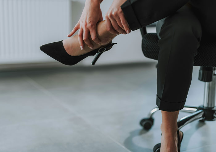 Person in black pants sitting on office chair, adjusting black high heel shoes with hands, in a modern workspace.
