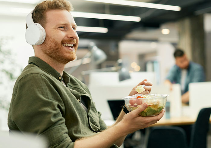 Man wearing headphones eating a salad in a modern office, representing job candidates asking mental questions to recruiters