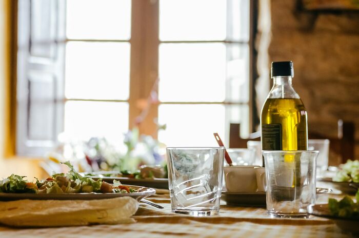 Rustic Greek village dining table with olive oil, fresh salad, and glassware near sunlit window. Rustic Greek village dining table with olive oil, fresh salad, and glassware near sunlit window.