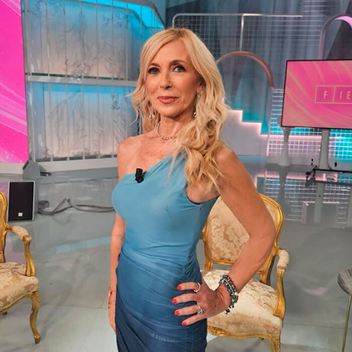 Woman in a blue dress posing indoors with blurred chairs and screens, related to Kate Middleton&rsquo;s facial scar rumors.