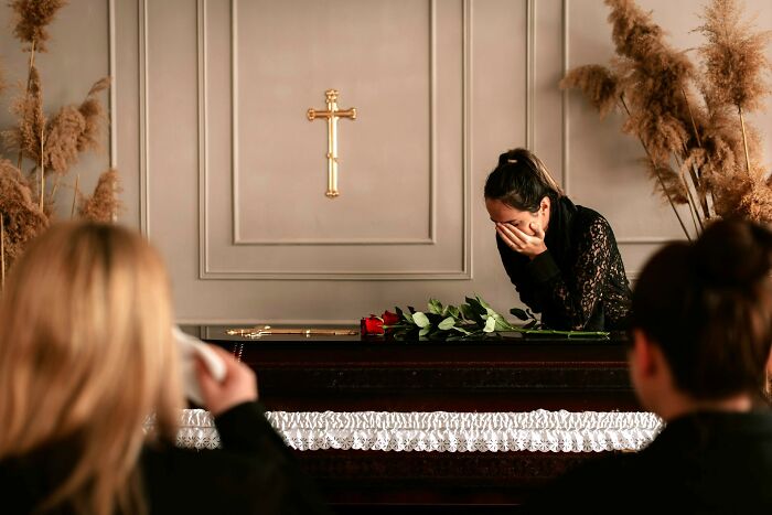 Woman grieving beside a coffin during a funeral service with a gold cross on the wall in a somber setting.