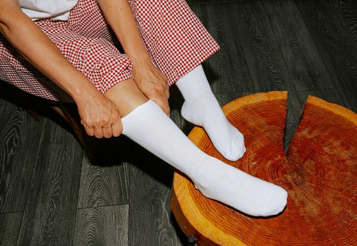 Person wearing white socks and red checkered pants, adjusting socks while sitting near a wooden stump on dark floor showing everyday things done wrong.