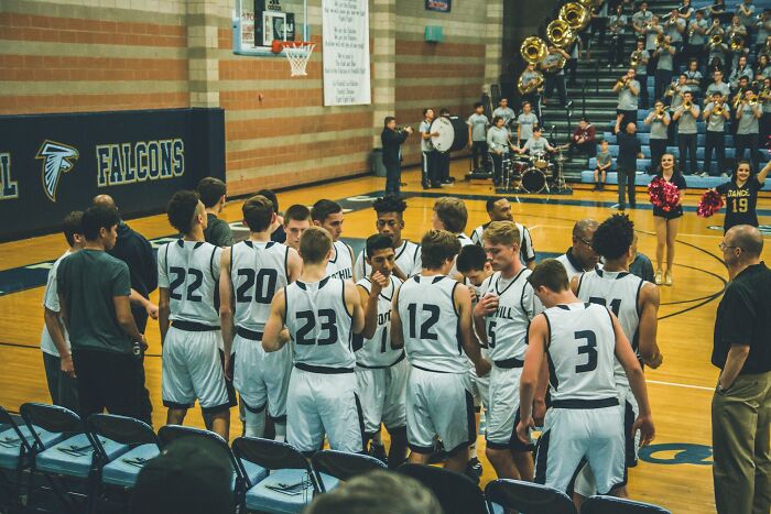 High school basketball team huddling during a game with cheerleaders and band in the background showing high school era vibes.