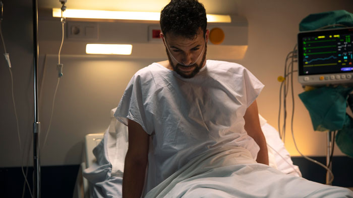 Man in hospital gown sitting on bed, appearing contemplative, with medical equipment in the background. Man in hospital gown sitting on bed, appearing contemplative, with medical equipment in the background.