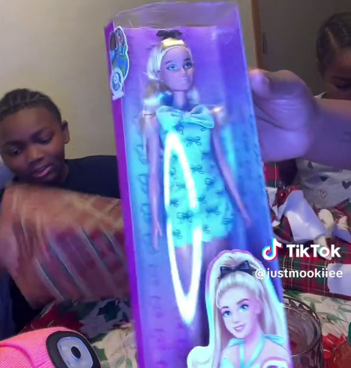 Child opening Angel Tree gifts, showing reaction to toy doll while mother films for TikTok video.