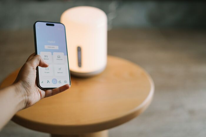 Hand holding smartphone with smart home app open, controlling a white smart lamp on a wooden table, showing disturbing facts concept.
