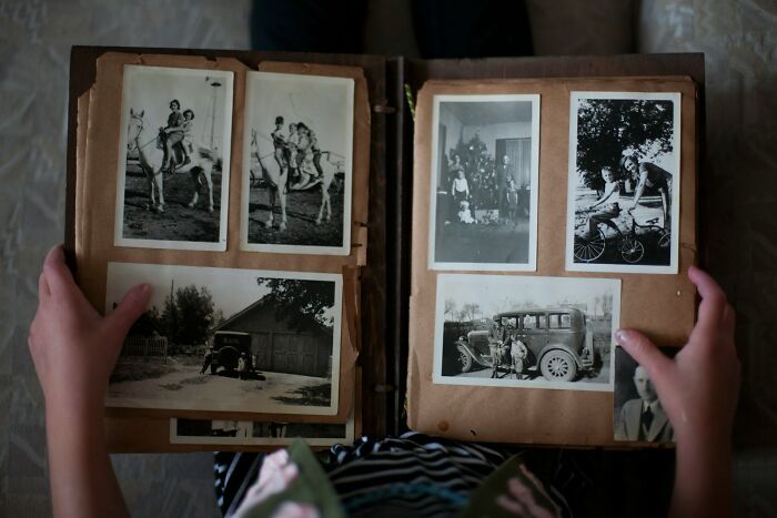 Hands holding a vintage photo album containing old black and white pictures of people and classic cars, unusual personal belongings.