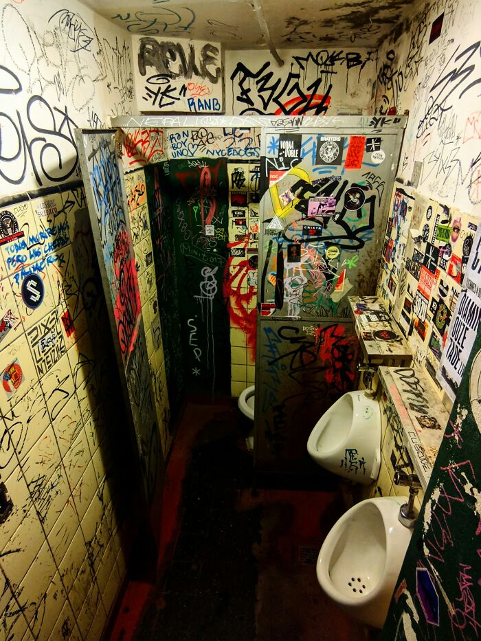 Graffiti-covered grimy bathroom stall with urinals and walls filled with sneaky acts of revenge stickers and tags.