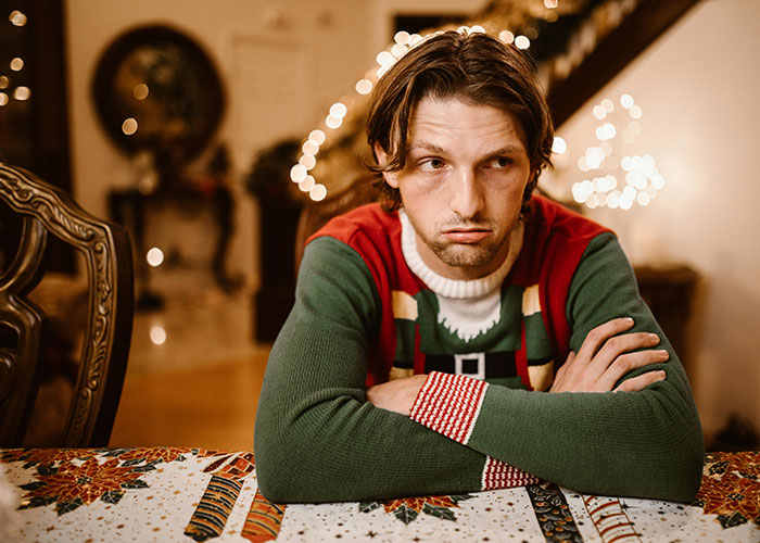 Young man in a Christmas sweater sitting at a dinner table looking upset during a family holiday gathering.