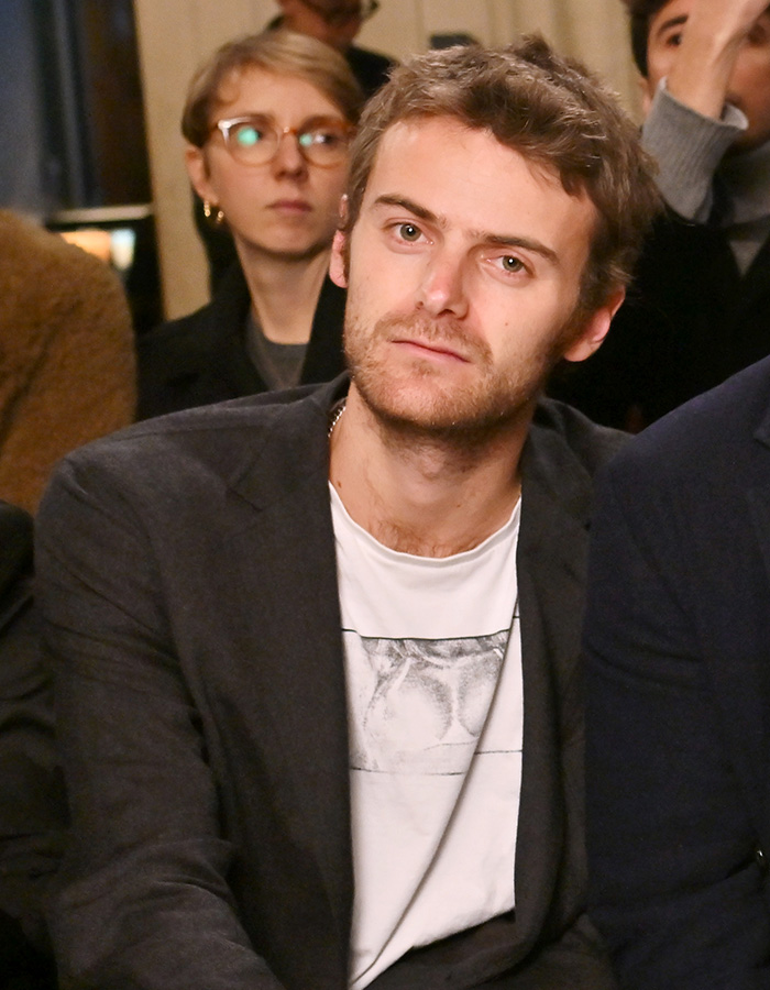 Young man with light brown hair and beard wearing a blazer and graphic t-shirt in a casual indoor setting. Young man with light brown hair and beard wearing a blazer and graphic t-shirt in a casual indoor setting.