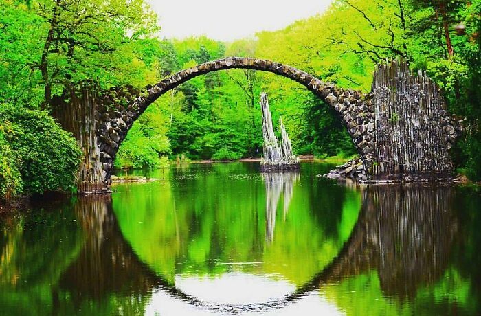 Natural stone arch bridge over calm water surrounded by lush green trees in a place that looks AI generated but is real.