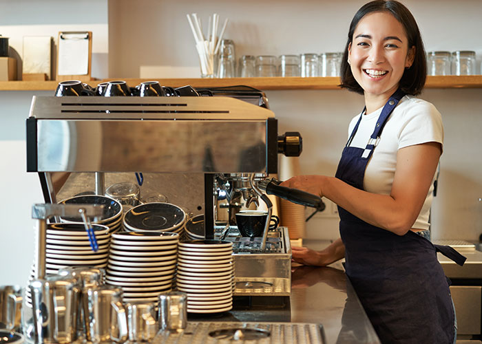 Coffee shop worker preparing espresso, smiling behind the counter with coffee machine and stacked cups in a busy cafe environment. Coffee shop worker preparing espresso, smiling behind the counter with coffee machine and stacked cups in a busy cafe environment.