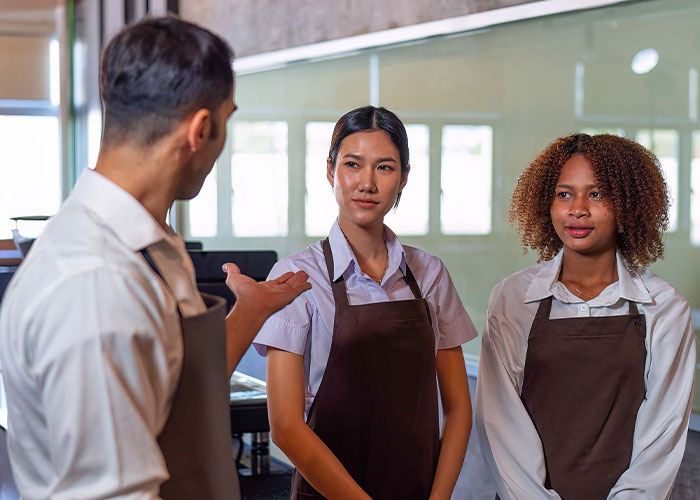 Coffee shop manager instructs workers on new rules while employees listen attentively during a team meeting. Coffee shop manager instructs workers on new rules while employees listen attentively during a team meeting.