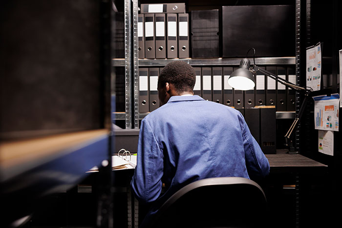 Man in blue shirt sits at dark desk, surrounded by binders and documents, pondering wedding and private investigator findings. Man in blue shirt sits at dark desk, surrounded by binders and documents, pondering wedding and private investigator findings.