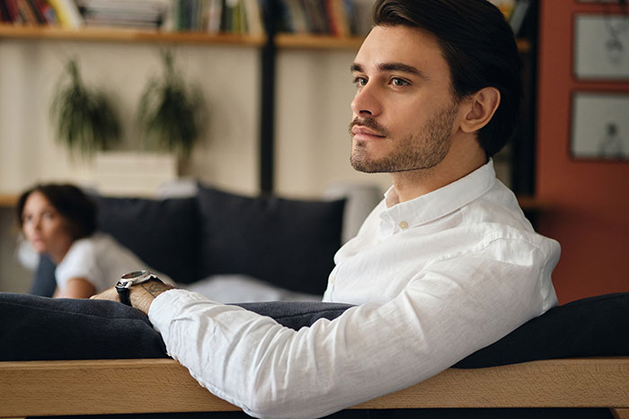 Man in white shirt with confident expression mansplaining to woman in casual home setting with blurred background.