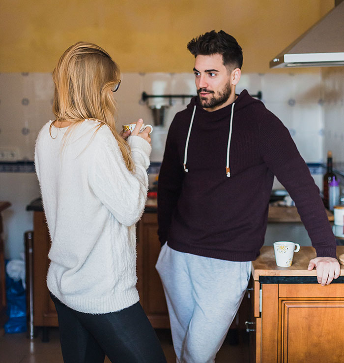A man mansplaining confidently to a woman in a cozy kitchen setting, both holding coffee cups during a casual conversation.