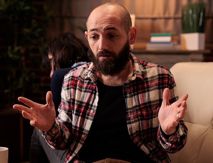 Bearded man in a plaid shirt mansplaining confidently to a woman in a casual home setting.