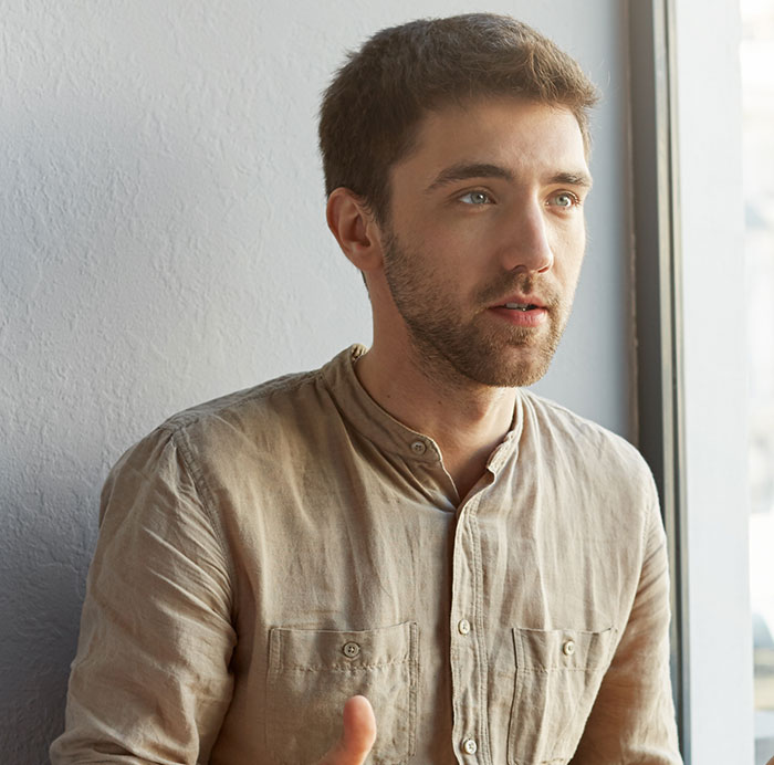 Young man mansplaining with confidence by a window, wearing a beige button-up shirt in a casual setting