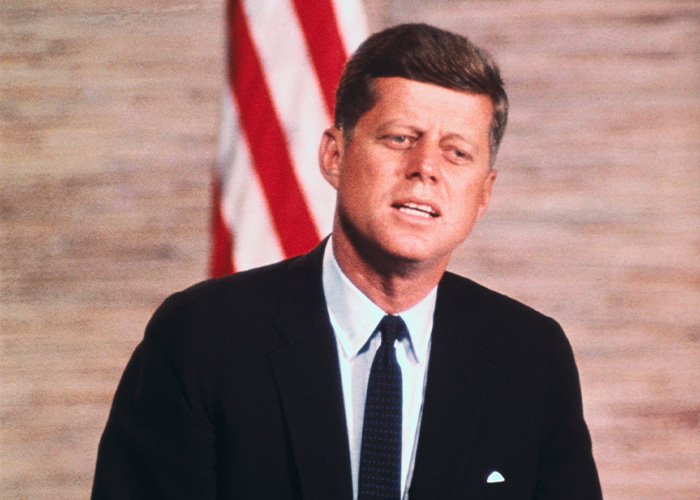 Man in a dark suit speaking in front of a blurred American flag, related to Kennedy Center renaming controversy online.